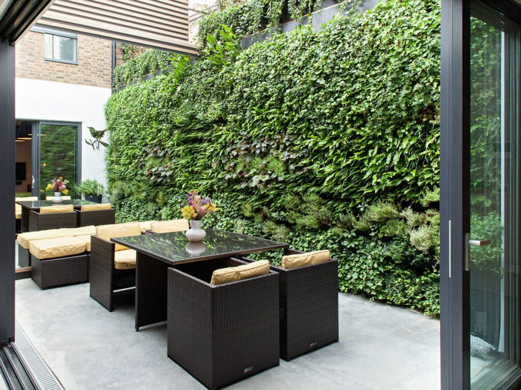 Vertical plant wall in the backyard of a house with chairs, and a vase placed on the table